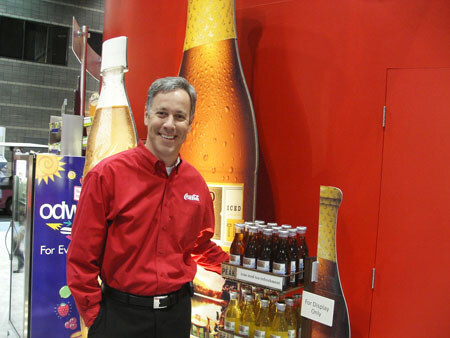 A smiling man in a red Coca-Cola shirt stands beside a large bottle graphic and a shelf of small bottles, next to an Odwalla banner, at a trade show booth.