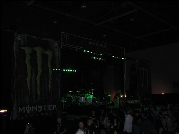 A dark hall with a stage lit by green lights and a drum kit. Large Monster Energy banners hang on both sides, and a crowd stands in the foreground.