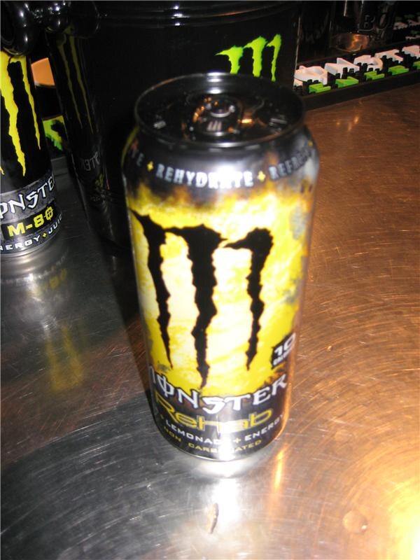 Close view of a yellow and black Monster Rehab lemonade energy can on a metal counter. A Monster M-80 can and other Monster Energy cans are in the background.
