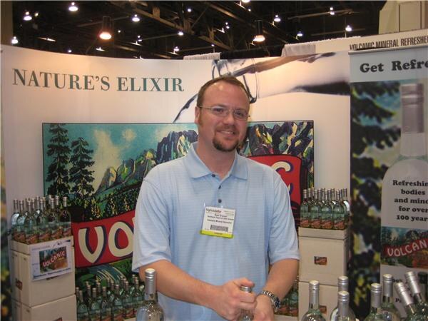 A smiling man with glasses and a goatee wearing a light blue polo and name badge stands at a Nature's Elixir booth with cases and bottles of Volcanic Mineral Refresher.