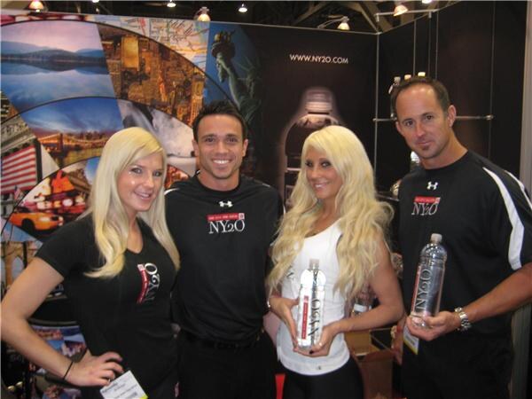Two women and two men pose in the NY2O booth. Two women hold tall NY2O bottles, and the men wear black Under Armour shirts with NY2O logos. A New York themed backdrop is behind them.