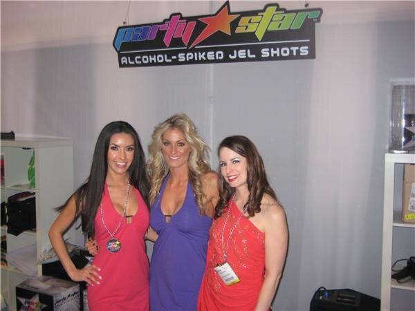 Three smiling women in red and purple dresses with lanyards stand together in front of a Party Star sign reading Alcohol-Spiked Jel Shots, with shelves on either side.