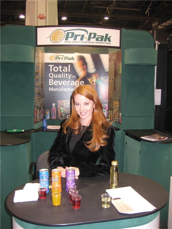 A smiling red-haired woman sits at a table in the green Pri-Pak booth. Switch cans and small cups of colored drinks are in front of her, and a banner reads Total Quality in Beverage Manufacturing.