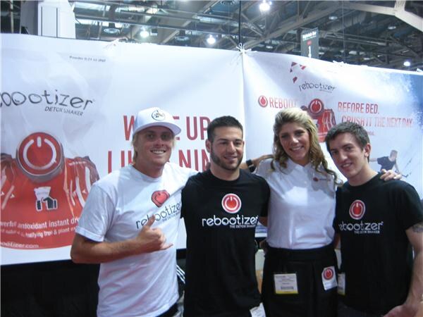Four smiling people, three men and a woman, stand in front of Rebootizer banners. Most wear Rebootizer shirts, and the woman wears a white shirt and a badge.