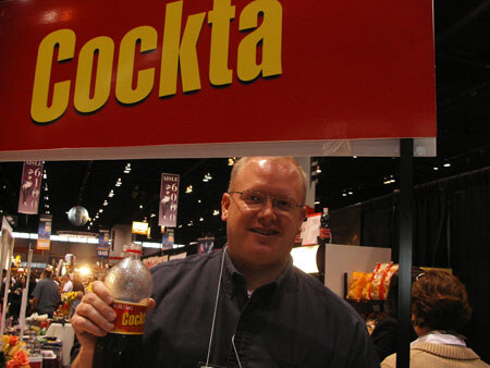 A smiling man with glasses holds a Cockta bottle under a red Cockta sign at a trade show, with other exhibits and attendees visible behind him.