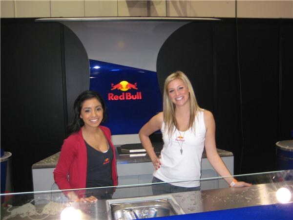 Two smiling women stand behind a counter in front of a blue Red Bull logo display; one wears a red cardigan and the other a white Red Bull tank top.