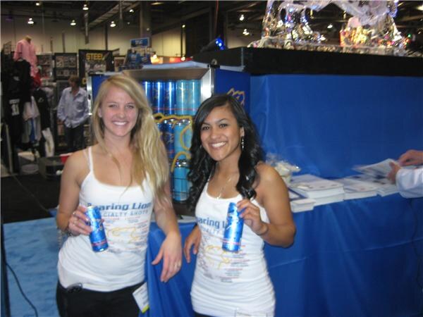 Two smiling women in white Roaring Lion Specialty Shots tank tops each hold a blue can beside a cooler stocked with cans, with a blue draped table behind them.