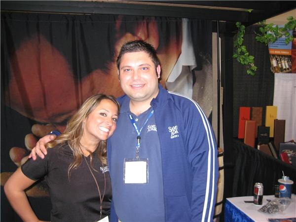 A smiling woman in a black Sam Adams shirt leans against a man in a blue Sam Adams jacket with a lanyard. A table with a Pepsi cup and a soda can sits at right.