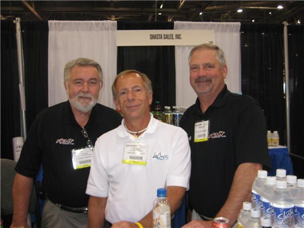 Three men stand behind a table at the Shasta Sales, Inc. booth. Two wear black polos and one a white polo with a LaCroix logo. Bottles of Shasta water are in front of them.