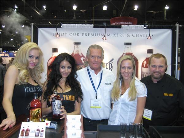 Five people stand behind a counter in front of a banner reading Enjoy Our Premium Mixers and Chasers. Two women hold bottles of Tamer mixer, and bottles and glasses sit on the counter.