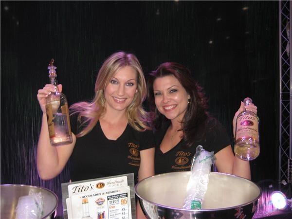 Two smiling women in black Tito's Handmade Vodka shirts each hold up a bottle of vodka behind a metal ice bucket, with an awards sign in front of them.