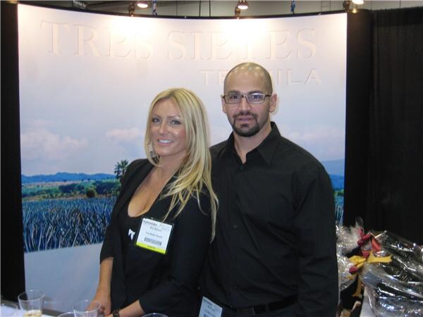 A blonde woman wearing a name badge and a bald bearded man with glasses in a black shirt stand together in front of a Tres Sietes Tequila backdrop showing an agave field.