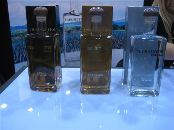 Three square Tres Sietes Tequila bottles labeled Anejo, Reposado and Silver stand on a glossy white display table in front of boxes and an agave field backdrop.