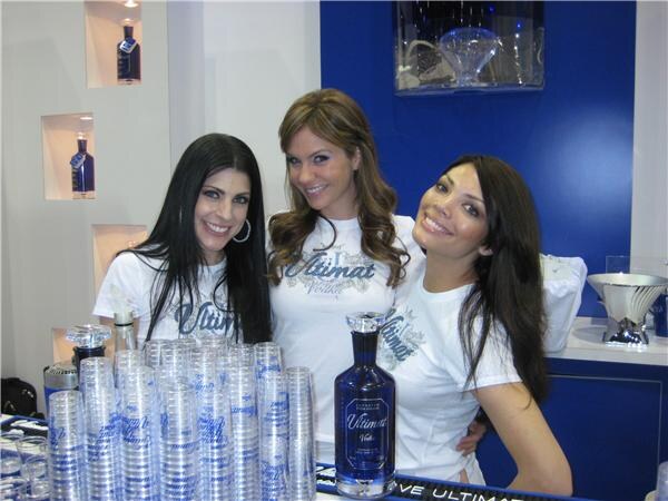 Three smiling women with long dark or brown hair in white Ultimat Vodka shirts stand behind a bar with stacks of plastic cups and an Ultimat bottle in front of a blue wall.