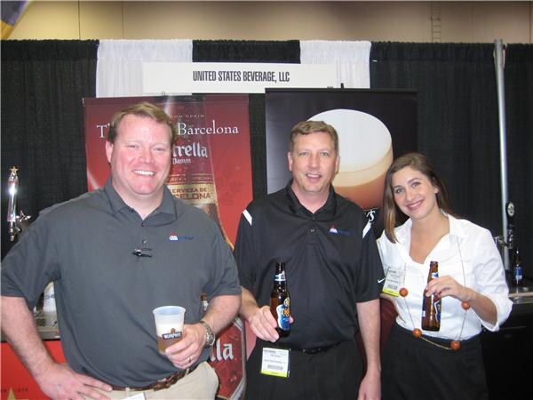 Two smiling men and a woman stand at a booth under a United States Beverage, LLC sign, with Estrella Damm banners behind them; two hold beer bottles and one holds a cup of beer.