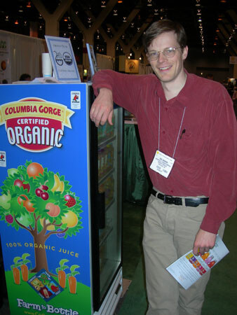 A smiling man with glasses and a name badge leans on a Columbia Gorge Certified Organic juice refrigerator, holding a brochure, at a trade show.