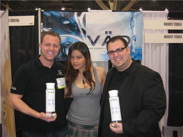 Two men, one in black polo and one in black jacket with glasses, hold white VÄD bottles on either side of a woman in a gray tank top, in front of a VÄD banner and Muchote Tequila signs.