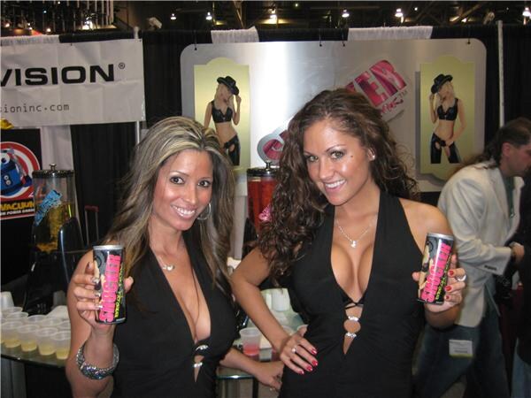 Two smiling women in black outfits each hold a can of energy drink in front of a Vision banner and display panels, with sample cups on the counter beside them.
