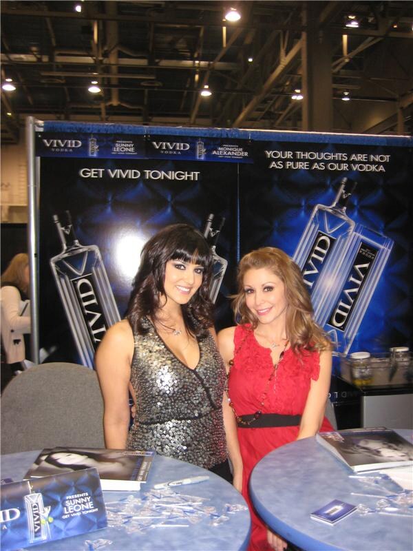 Two smiling women, one in a sequined gray top and one in a red dress, stand behind blue tables at a Vivid Vodka booth with large bottle banners reading Get Vivid Tonight.