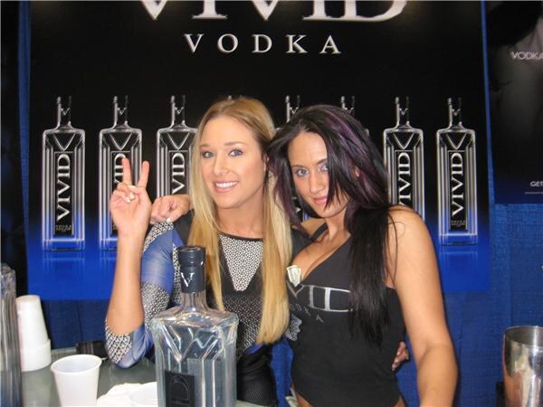 Two women pose behind a counter holding a Vivid Vodka bottle, one blonde making a peace sign and one with long dark hair, in front of a banner of Vivid Vodka bottles.