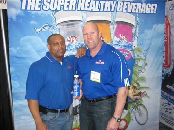 Two men in blue polo shirts stand together in front of a VPX banner reading The Super Healthy Beverage, one holding a small bottle; Redline is shown on the shirts and signage.