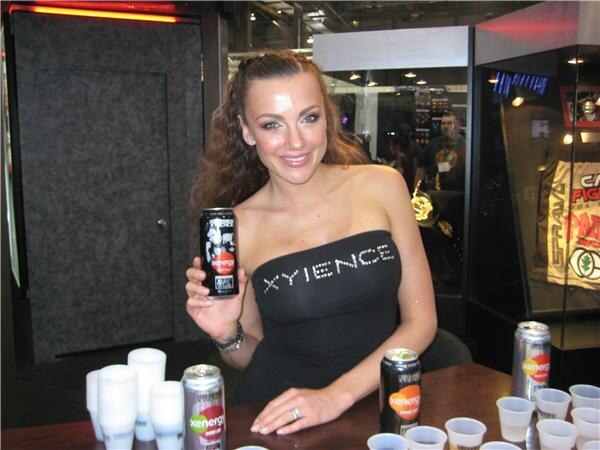 A woman with long wavy brown hair in a black strapless top reading Xyience holds a black Xenergy can at a table with more Xenergy cans and small cups.
