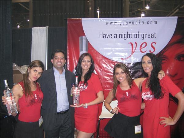 A man in a dark suit stands among four women in red YES Vodka outfits, several holding vodka bottles, in front of a red banner reading "Have a night of great yes" and yesvodka.com.