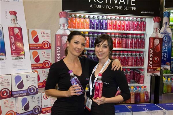 Two women in black tops hold bottles of Activate drinks in front of shelves of bottles and stacked Activate Workout boxes beneath a sign reading "Activate, a turn for the better".