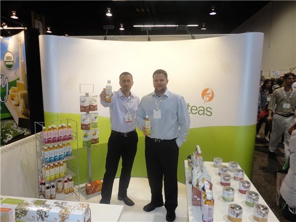 Two men in light blue shirts and badges hold up bottled teas in front of a curved white and green Adagio Teas display, with shelves of bottles and tins beside them.