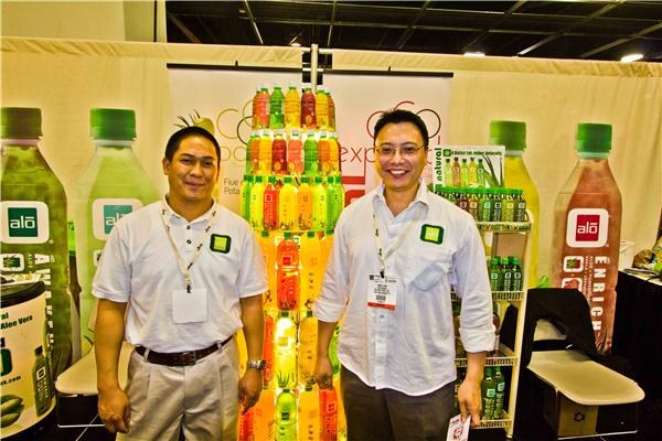 Two men in white shirts with lanyards stand smiling in front of a stack of colorful alo aloe drink bottles and large alo bottle graphics.