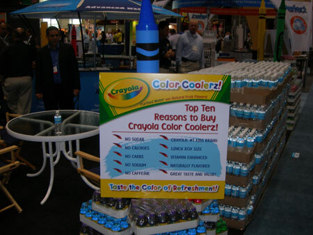 A display of stacked Crayola Color Coolerz water bottles with a sign listing ten reasons to buy, beneath a giant crayon, with attendees in the background.
