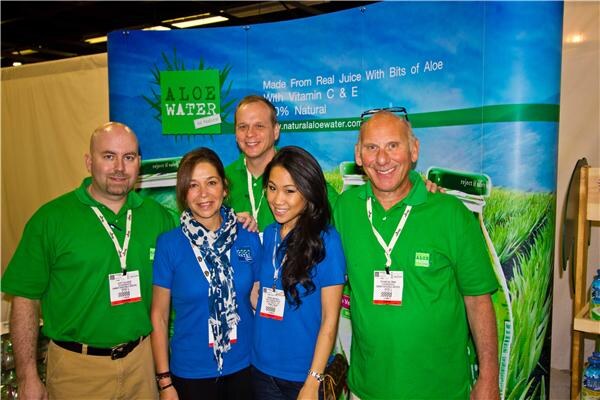 Four people in green and blue shirts with badges stand together, with a fifth person behind them, in front of an Aloe Water banner showing a grassy field and the text "Made from real juice".