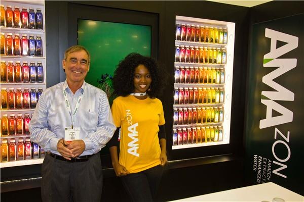 A smiling man in a striped shirt and a woman in a yellow Amazon t-shirt stand between lit shelves of colorful bottles, beside a black sign reading Amazon enhanced water.