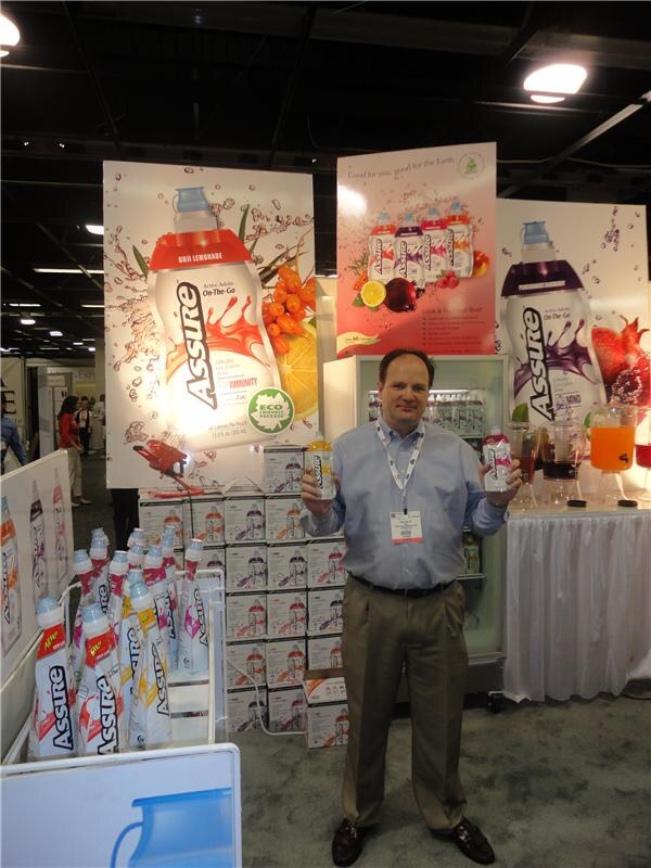 A man in a blue shirt and khakis holds up two Assure drink containers in a booth with large Assure banners, stacked cases, a cooler and a table of juice dispensers.