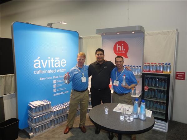 Three men in blue and black shirts stand with arms around each other in a booth with a blue ávitāe caffeinated water banner, a round table with bottles, and a bottle cooler.