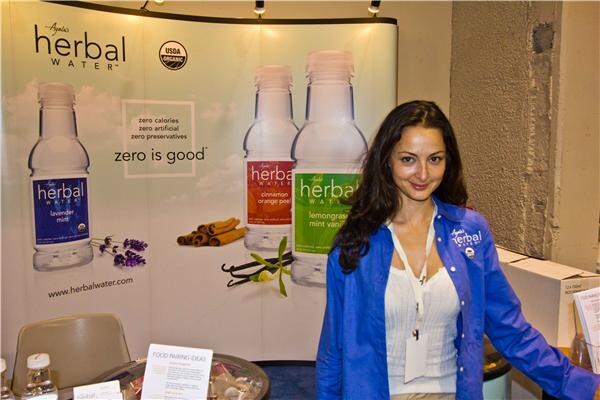 A woman with long dark hair in a blue Ayala's Herbal Water jacket stands in front of a banner showing herbal water bottles and the words "zero is good".