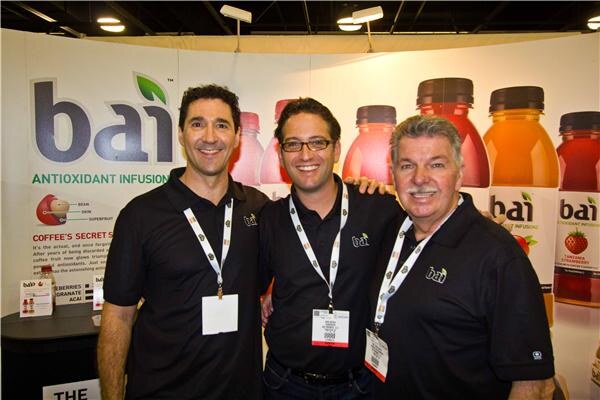 Three men in black bai polo shirts and lanyards smile together in front of a bai antioxidant infusion banner and colorful bottles.