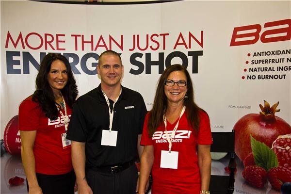 Two women in red BAZI Energy Shots shirts and a man in a black shirt stand together in front of a banner reading "More than just an energy shot" with pomegranate and raspberry images.