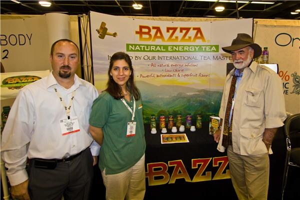 Three people stand in front of a Bazza Natural Energy Tea booth; a man in a white shirt, a woman in a green shirt and a bearded man in a hat. Tea bottles sit on the table behind them.