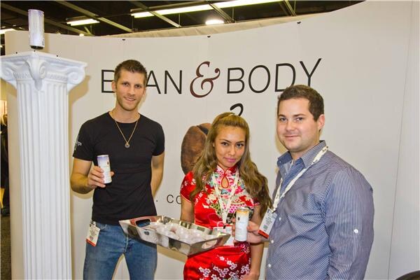 Two men and a woman in a red patterned dress stand at a Bean & Body booth, holding small cans and a tray of samples beside a white decorative column.
