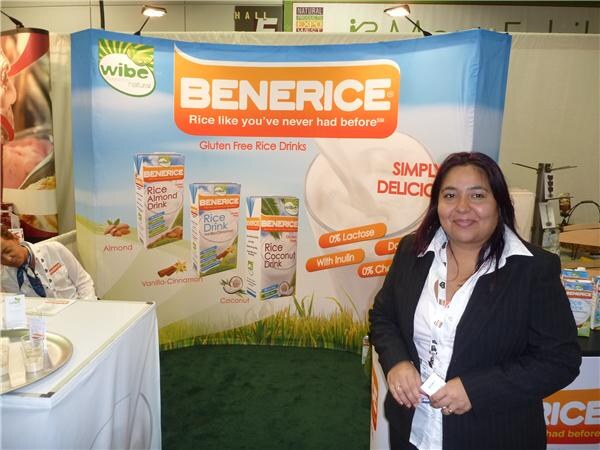 A woman in a dark jacket stands in front of a Benerice booth by Wibe Natural, with banners for gluten free rice drinks in almond, vanilla-cinnamon and coconut flavors.