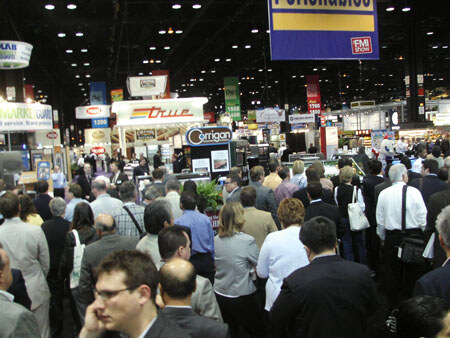A dense crowd of attendees fills an aisle of the exhibit hall, with overhead signs, a FMI Show banner and a Corrigan booth sign in the background.