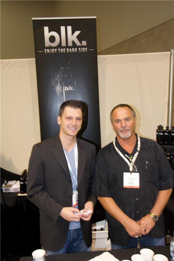 Two men stand smiling in front of a black blk. banner reading "Enjoy the dark side," with dark bottles of blk. water on the table beside them.