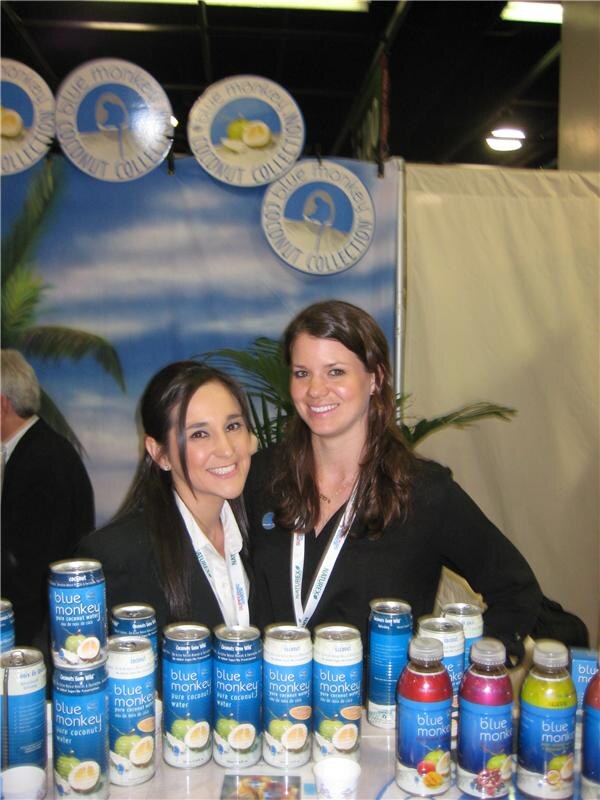 Two smiling women stand behind a table of Blue Monkey pure coconut water cans and bottles, with a Blue Monkey Coconut Collection beach backdrop behind them.