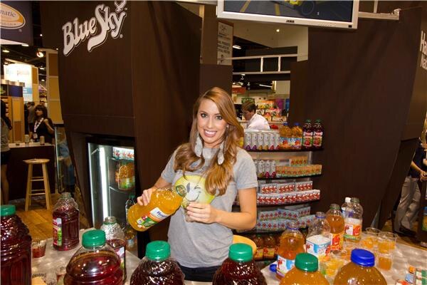 A smiling woman in a gray T-shirt pours a drink from a bottle at the Blue Sky booth, with bottles of juice and soda on the counter and shelves of product behind her.