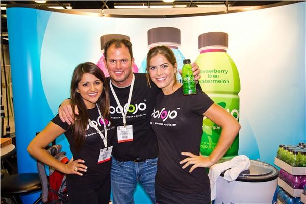 Two women and a man in black Bojo T-shirts pose together in front of large Bojo bottle graphics; one woman holds a green bottle.