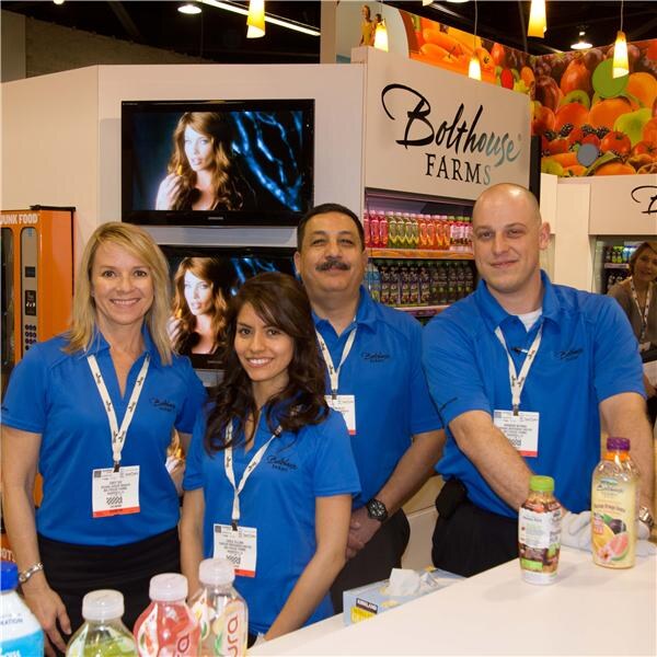 Four people in blue Bolthouse Farms polo shirts and badges stand behind a counter of bottled drinks, with televisions and a Bolthouse Farms sign behind them.