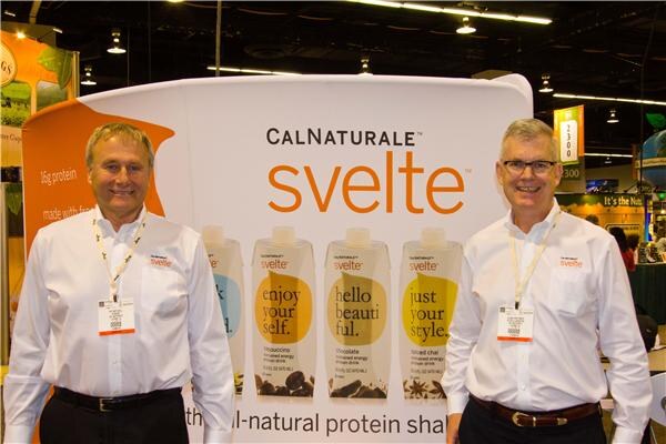 Two men in white shirts with CalNaturale Svelte logos stand in front of a display of Svelte protein shake cartons in several flavors.