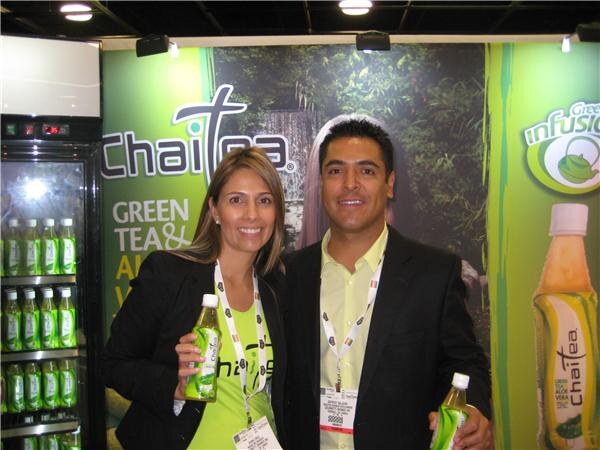 A smiling woman and man in dark jackets and lanyards stand in front of a green ChaiTea banner, each holding a bottle of ChaiTea green tea. A refrigerated case of bottles is at left.