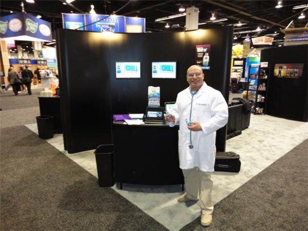 A man in a white lab coat with a stethoscope stands holding a small bottle beside a black display counter with two Chill screens in a large exhibition hall.